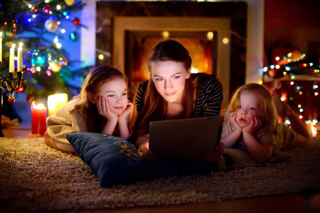 A mother and her children watching a Christmas movie on a tablet near the Christmas tree.
