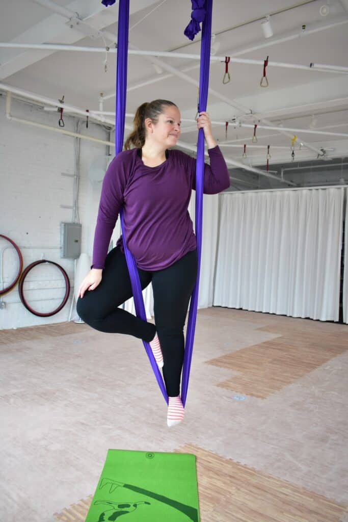 A woman doing a tree pose in an aerial silk above a yoga mat.