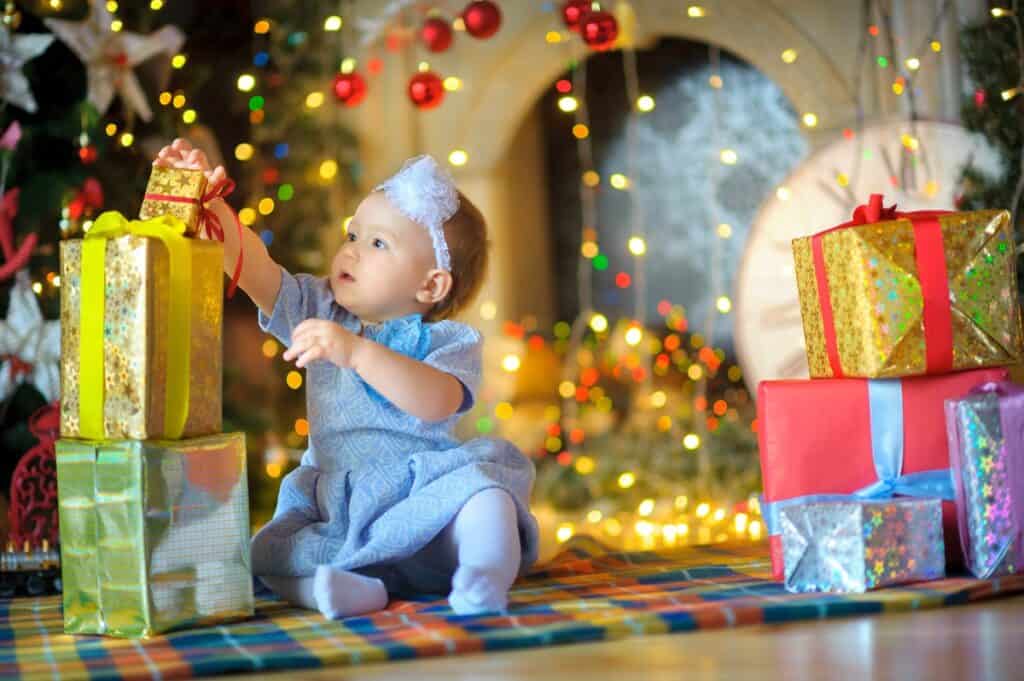 A young child sitting on the floor next to Christmas gifts and a Christmas tree.